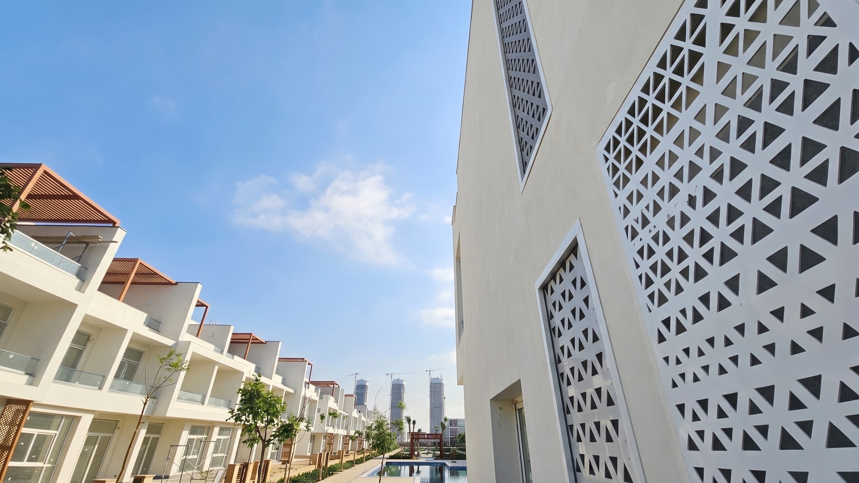 A white townhouse facade with triangular perforated screen panels, a landscaped pool spine and towers under construction beyond.